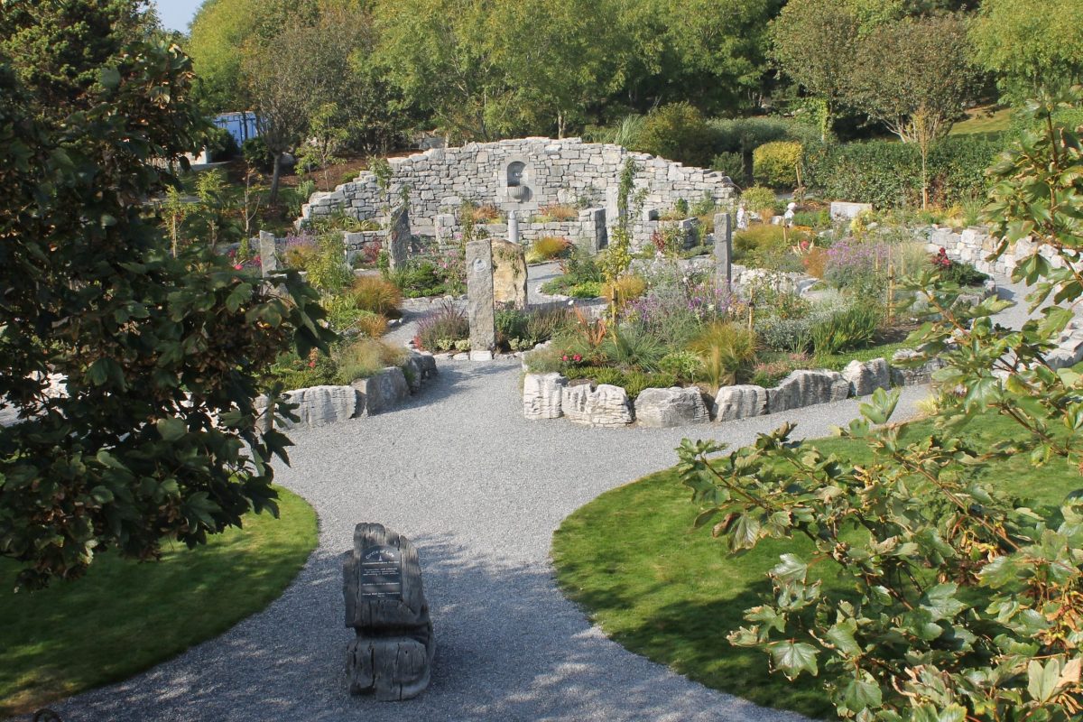 Landscaped memorial garden near Salthill, Galway, featuring winding gravel paths, native planting, stone markers, and limestone walls surrounded by mature trees. The peaceful green space offers a quiet setting for reflection and gentle walks close to the seafront.