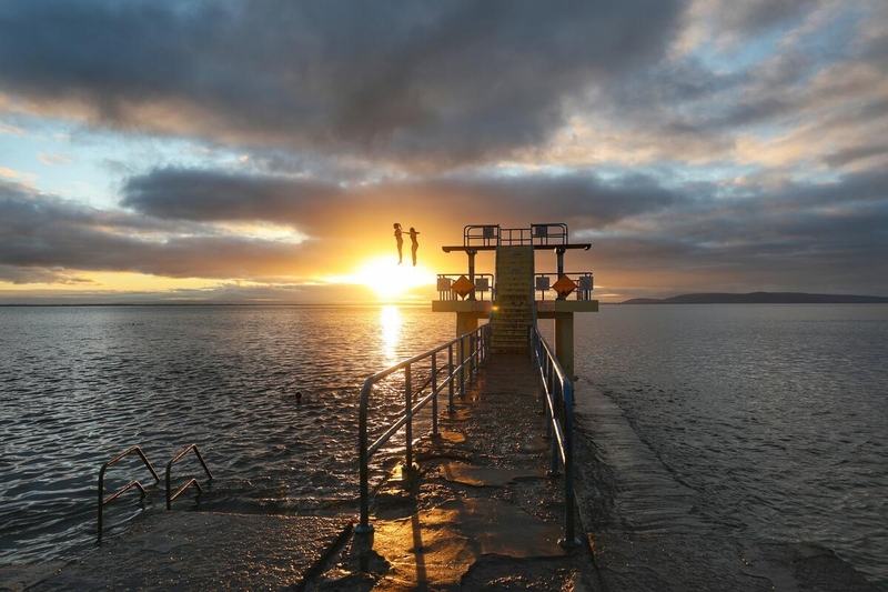 Blackrock Diving Tower in Salthill, Galway, silhouetted against a sunset over Galway Bay, with swimmers and calm sea waters surrounding the landmark.